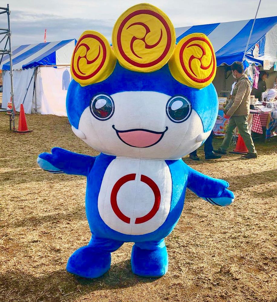 A blue catfish mascot with arms and legs and wearing yellow Tomoe swirls on a headband poses in a field, in front of some stripy tents.
