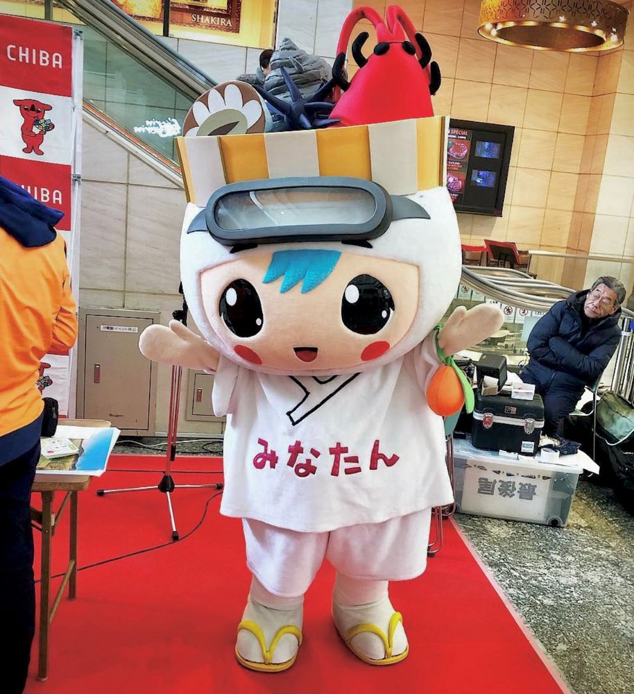 A mascot in an old-fashioned white diving outfit and  with a bowl of seafood on her head stands on a red carpet.