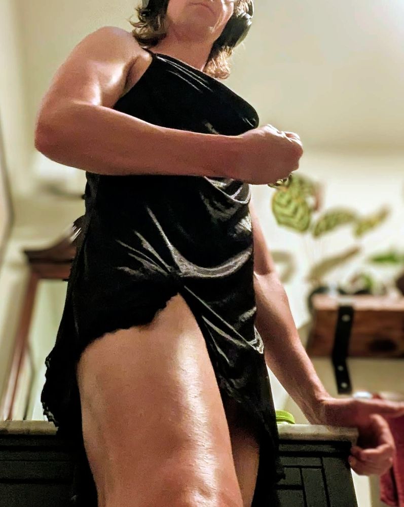 Necluda in a black slip dress flexing and looking down at you.