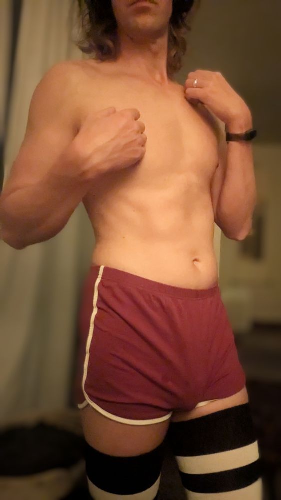 A person in burgundy short shorts and black and white thigh high stockings posing with their tummy exposed. 