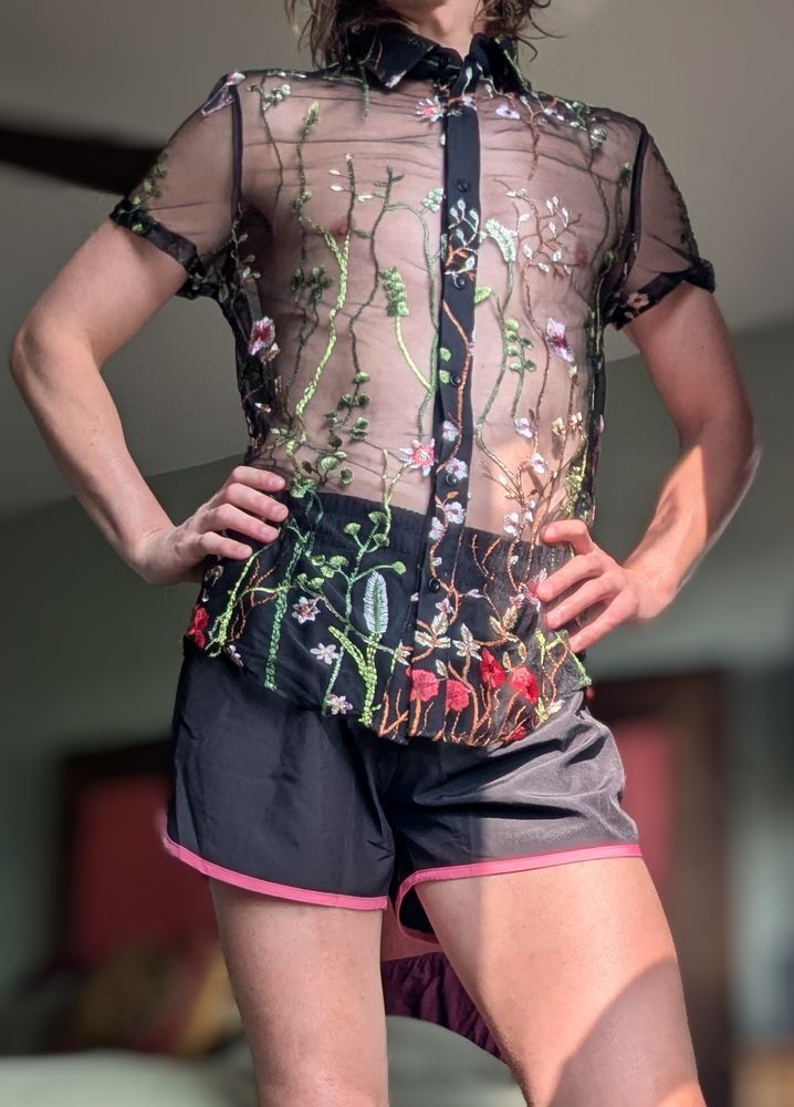A femboy with their hands on their hips wearing black and pink shorts and a mesh black see thru top with flowers. 