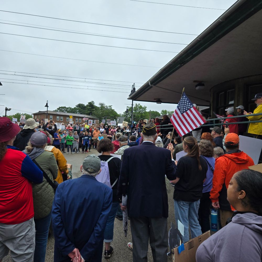 Crowd of people by the Elkins Park train station. In the center is a tall elderly white man with his American Legion cap and jacket, holding an American flag.