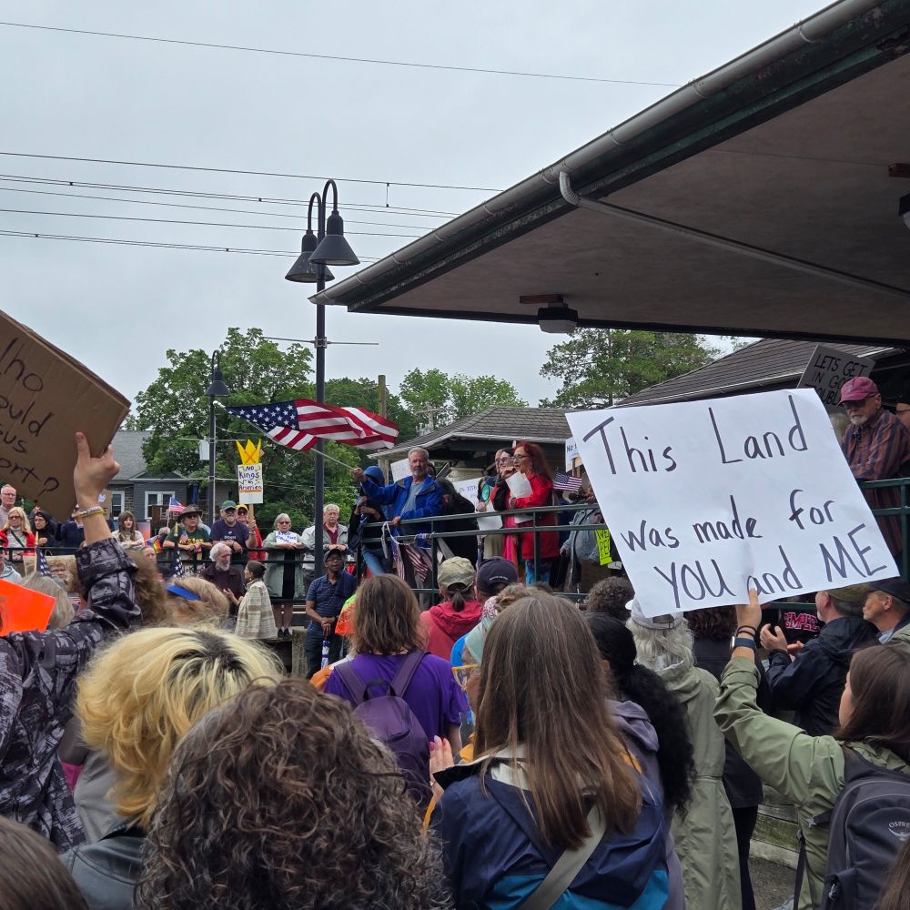 Crowd around train station. A man waves the American flag. A sign says This land was made for YOU and ME.