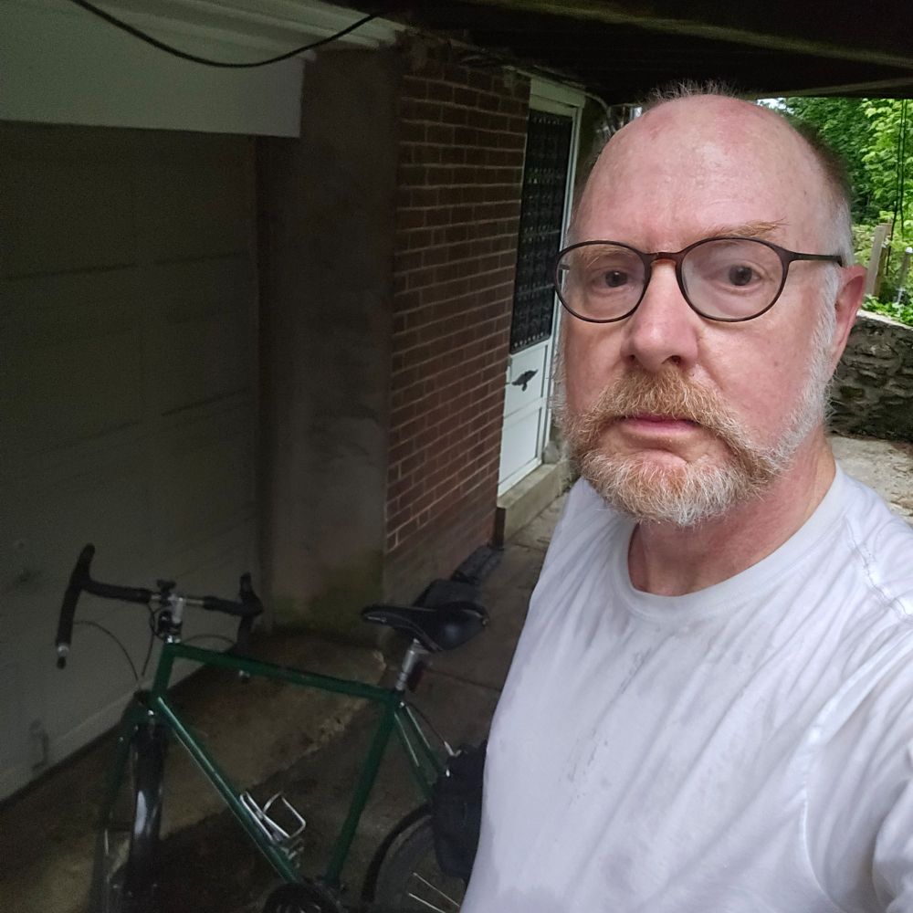 Tired, bald white man with a gray and red beard looks at the camera. His spectacles are steamed up. His white t-shirt is drenched with sweat. In the background is a green bicycle with black saddle and black dropped handlebars. HAPPY BIKE TO WORK DAY!