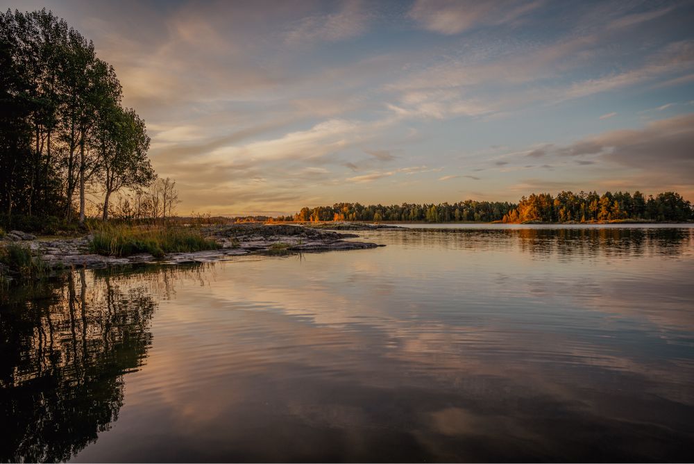 A serene view of Lake Vänern in Sweden during sunset. The sky is painted with soft hues of orange and pink, reflecting beautifully on the calm water. Trees line the rocky shoreline on the left, their silhouettes mirrored in the lake. The distant horizon features a mix of lush greenery and golden autumn foliage, creating a tranquil and picturesque landscape.