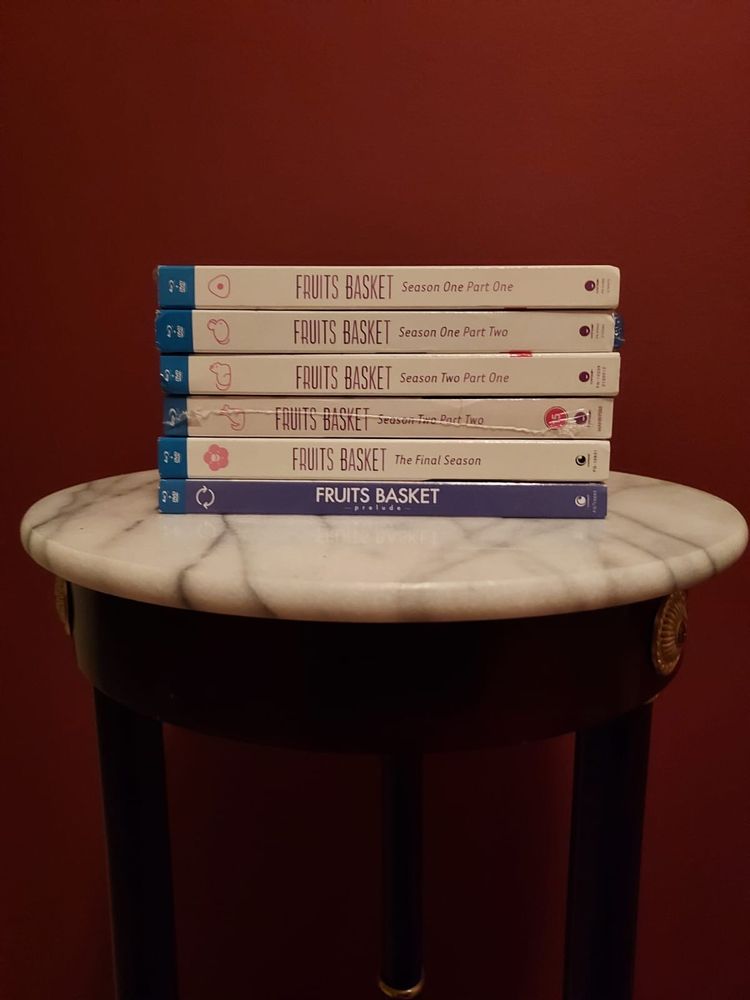 Photo of seven Fruits Basket Blu-Ray cases stacked neatly on a round marble-topped table against a deep red wall. Titles include all three seasons of the Fruits Basket anime and the Prelude movie.