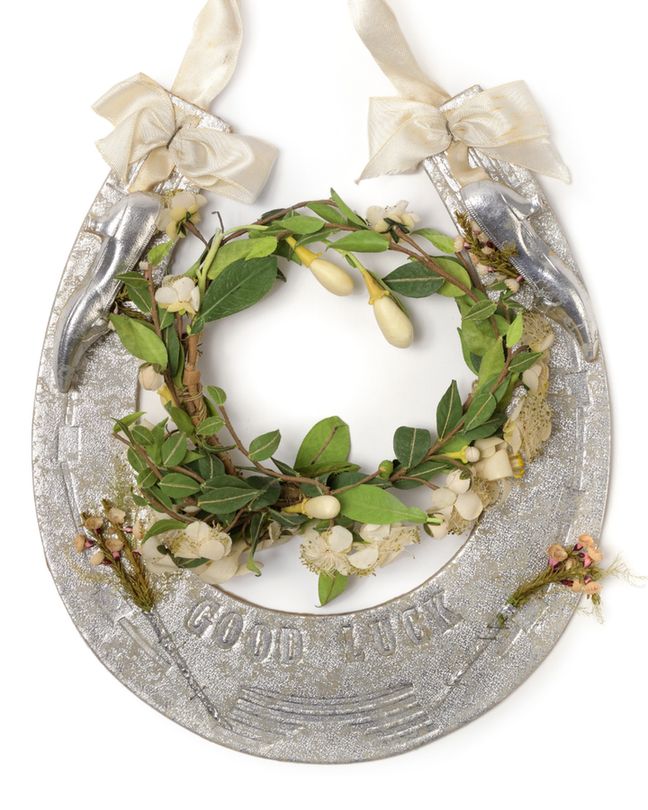 Close view of a silver‑coloured decorative horseshoe with “GOOD LUCK” embossed along the curve. Two cream ribbon bows sit at the top corners, each beside a small hanging silver shoe. A wreath of artificial green leaves and pale flowers fills the centre opening. Small bunches of artificial flowers are attached near the lower edge. Studio white background.