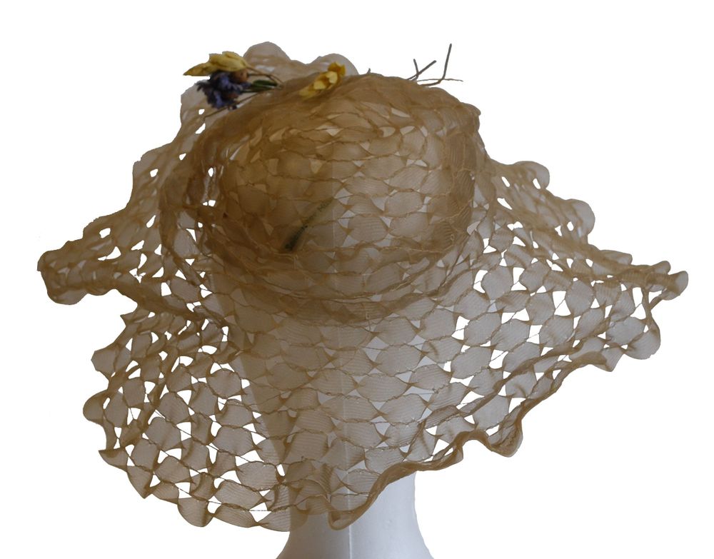 Light brown or beige wide-brimmed hat made from a sheer, open-weave fabric, creating a delicate lattice pattern throughout the crown and brim. The brim is undulating and floppy, contributing to a soft, flowing appearance. A small floral decoration with yellow and purple flowers and thin twigs adorns one side of the crown. The hat is displayed on a white mannequin head against a plain background.
