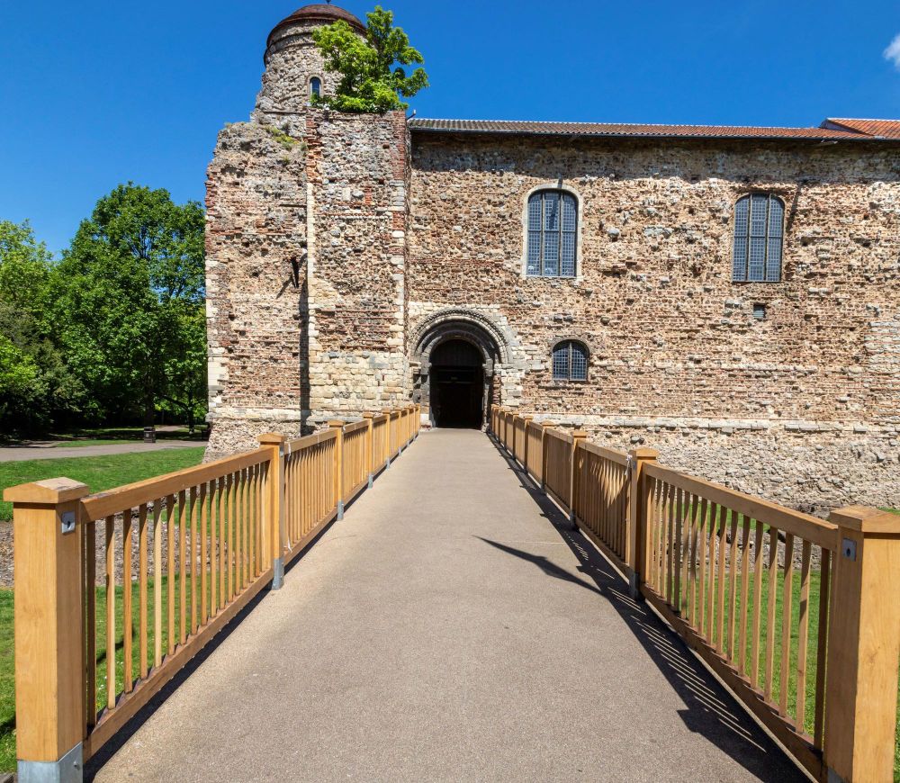 Colchester Castle, a historic stone building with a rugged, weathered exterior, likely a castle or medieval structure, stands beneath a bright blue sky. The building features arched windows with leaded panes and a prominent arched entrance. A small round tower on the left, partially overgrown with greenery, hints at the building's age and past fortifications. A long, wooden-railed walkway leads directly to the entrance, creating a sense of approach and inviting visitors to explore. The surrounding grounds are landscaped with well-kept green lawns and trees, adding a contrast between the building’s ancient stonework and the vibrant, clear day.