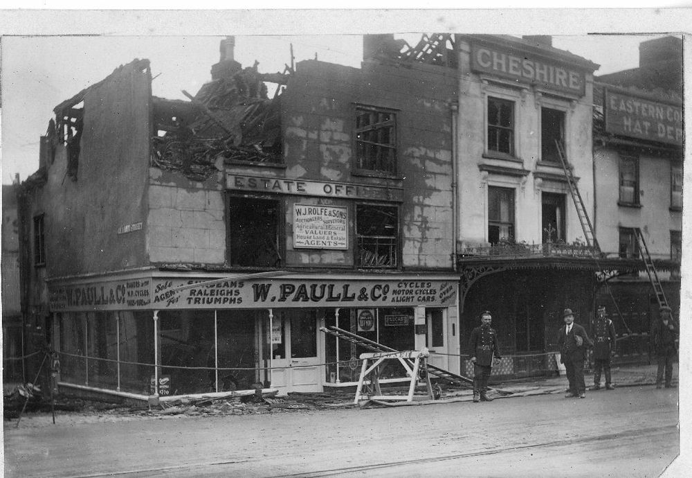 Black-and-white street scene showing a busy street corner after a shop fire, with fire hoses on the ground, a fire engine, ladders against a soot-stained shopfront, broken windows and a crowd of onlookers lining the pavement.