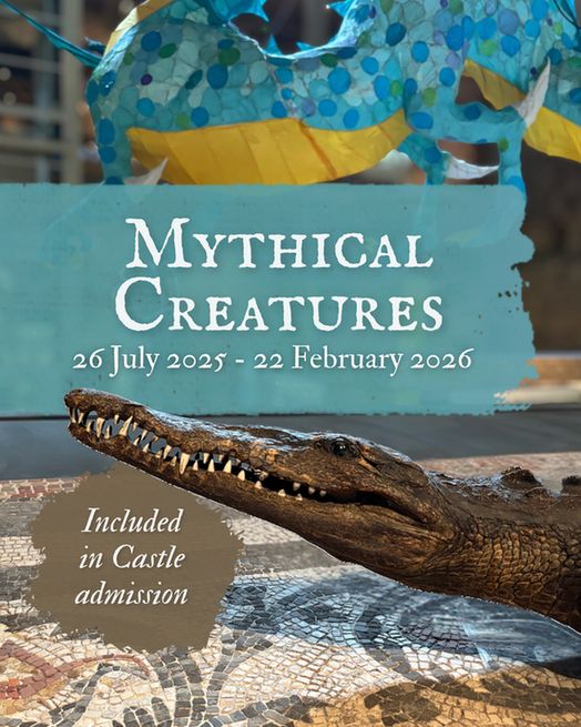 A promotional image for the Mythical Creatures exhibition at Colchester Castle, showing a crocodile model lying on a Roman mosaic floor. Above it, a blue and yellow paper dragon is suspended in the air. Text on the image reads: “Mythical Creatures, 26 July 2025 – 22 February 2026. Included in Castle admission.”