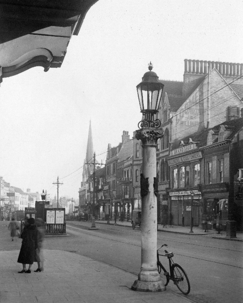 Black and white photograph of a bustling high street from the early to mid-20th century. A tall ornate gas lamp stands in the foreground with a bicycle leaning against its base. On the right, multi-storey buildings house various businesses, including "Brand Bros" tailors and costumiers. Overhead tram lines and tracks embedded in the road suggest active tram services. People in period clothing walk along the pavement, with a pair of women conversing in the left foreground. In the background, a church spire rises above the rooftops, partially obscured by haze.