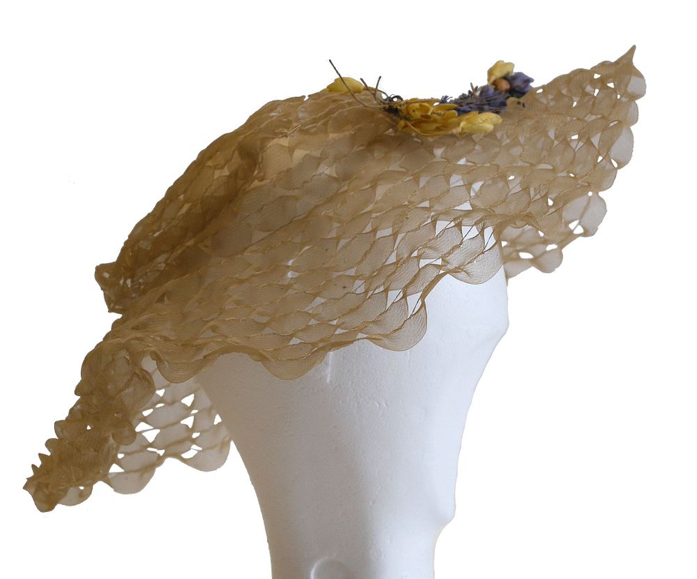 Side view of a light brown, open-weave hat with a wide, wavy brim and translucent lattice texture. The crown is decorated with artificial flowers in yellow and purple, along with small twig-like elements, placed slightly forward on the hat. The brim curves downward at the back and sides, adding to the delicate, airy appearance. Displayed on a white mannequin head, the hat conveys a vintage or romantic style.