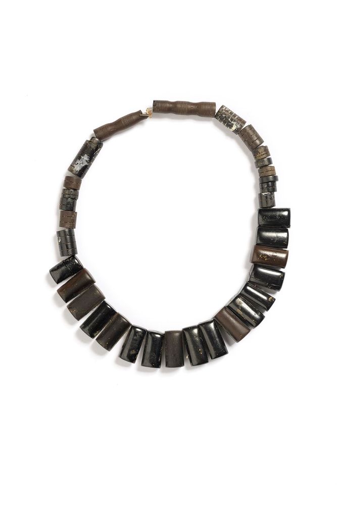 Armlet composed of large, tapered jet beads arranged in a semi-circle, with smaller cylindrical and disc-shaped beads forming the upper section. The jet beads are polished to a smooth, glossy finish in shades of deep black and dark brown, some showing natural inclusions or speckling. The smaller beads vary in texture and colour, including matte and slightly rougher surfaces. The armlet is displayed on a plain white background.
