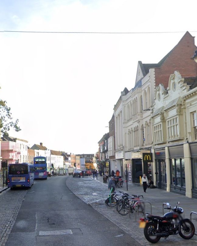 Busy town centre street lined with historic buildings featuring ornate façades. On the right, a McDonald's restaurant occupies part of a cream-coloured building with decorative mouldings. Several bicycles are parked at metal racks nearby, alongside a black motorcycle. Pedestrians walk along the pavement, and two double-decker buses are seen in the left lane, heading down the road. The sky is clear, and the atmosphere is lively with a mix of transport and foot traffic.