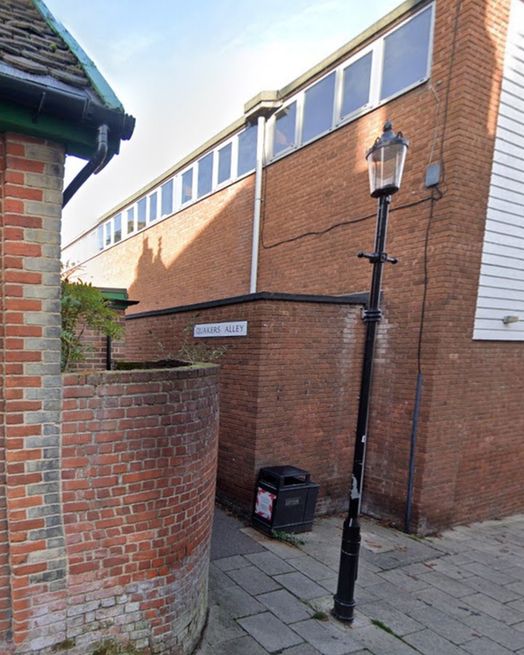 Modern colour photo of the same alleyway with updated pavement, signage reading "Quakers Alley," and the same curved brick wall now topped with plants.