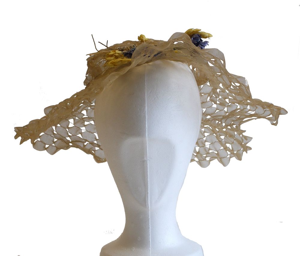 Front view of a light brown, sheer wide-brimmed hat with an open lattice design, displayed on a white mannequin head. The brim curves gently around the face, creating a soft, airy silhouette. Small artificial flowers in yellow and purple, along with thin twig-like accents, embellish the crown, slightly offset to one side. The hat appears lightweight and decorative, suitable for warm weather or formal occasions.