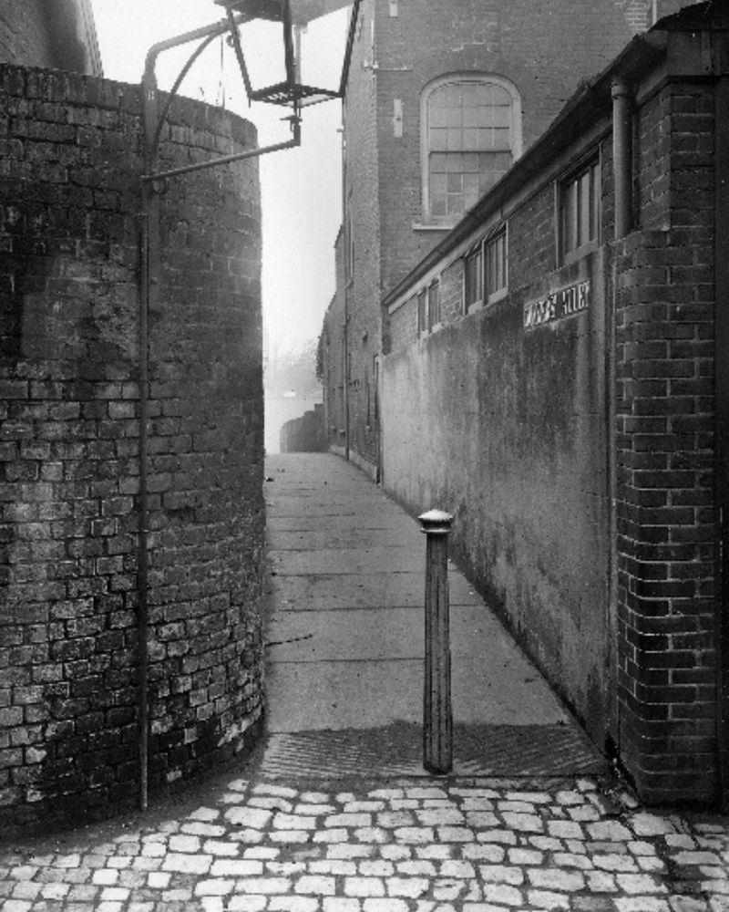 Black-and-white photo of a narrow alleyway bordered by tall brick walls and a single bollard. A curved corner wall and a large gas lamp overhang the entrance. The paving is cobbled and the alley appears misty in the distance.