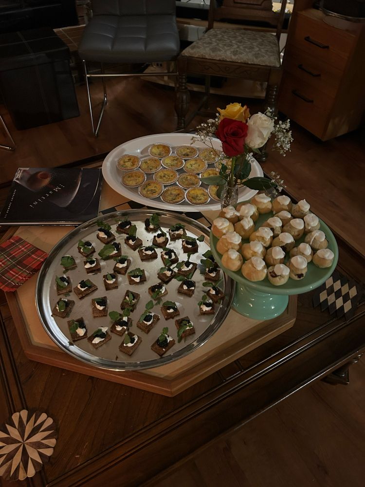 A tray of caviar on rye, cream puffs, and mini quiches, on a coffee table with a small bouquet of roses