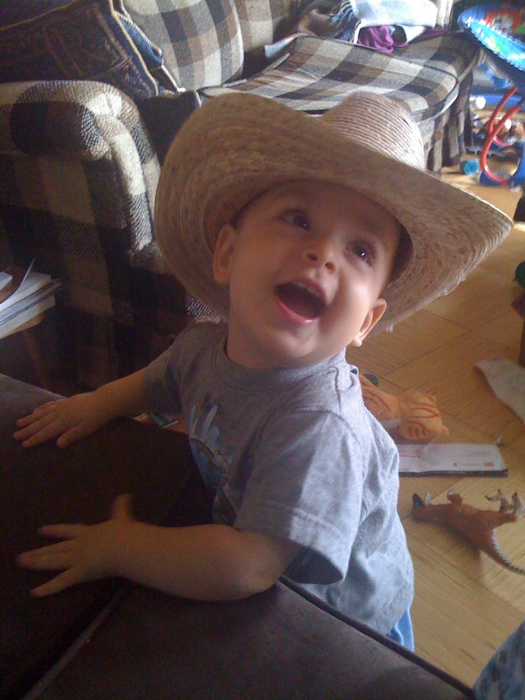 A baby standing by a couch wearing a straw hat and laughing