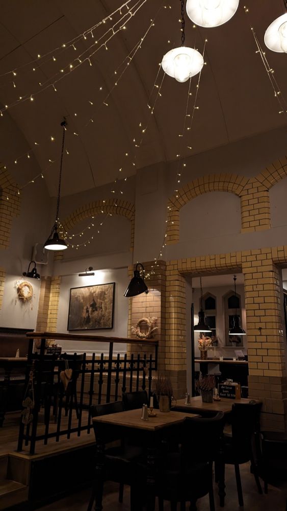 Zu sehen ist der Innenraum eines Restaurants. Im Vordergrund zwei leere Tische, dahinter eine leichte Erhöhung, die über zwei Stufen erreicht werden kann. Auch hier steht ein leerer Tisch. Ein schwarzes Geländer dient als zusätzliche räumliche Trennung.
Die Wände des Raumes sind grau. Gelbe Backsteinen sind als Zierde angebracht und wie Torbögen platziert. Im Hintergrund sieht man die Bar der Location.
Von der Decke hängen schwarze, trichterförmige Lampen und Lichterketten. Diese sind durch den ganzen Raum gespannt, wie Girlanden. Ihr Licht ist warm und gemütlich.