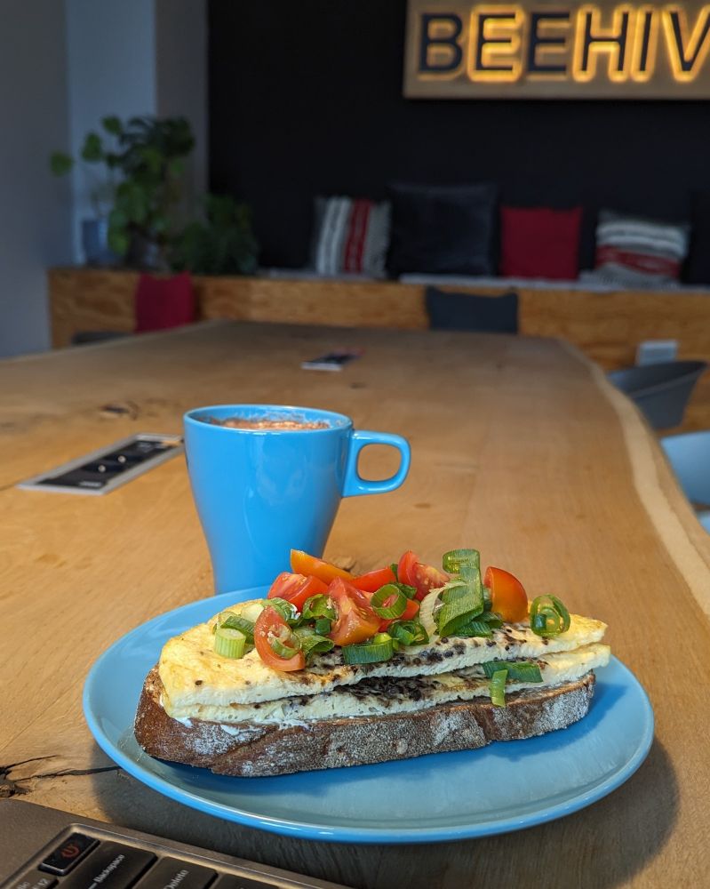 Ein hellblauer Teller und eine hellblaue Tasse auf einem langen Holztisch. Auf dem Teller liegt eine Scheibe Brot, darauf Omelett, geschnittene Frühlingszwiebeln und geschnittene Tomaten.
Am linken unteren Rand des Bildes ist noch eine Laptop-Tastatur zu erkennen. Im Hintergrund eine Sitzecke mit Kissen und an der Wand ein beleuchteter Schriftzug mit dem Namen des Co-Working-Spaces: Beehive