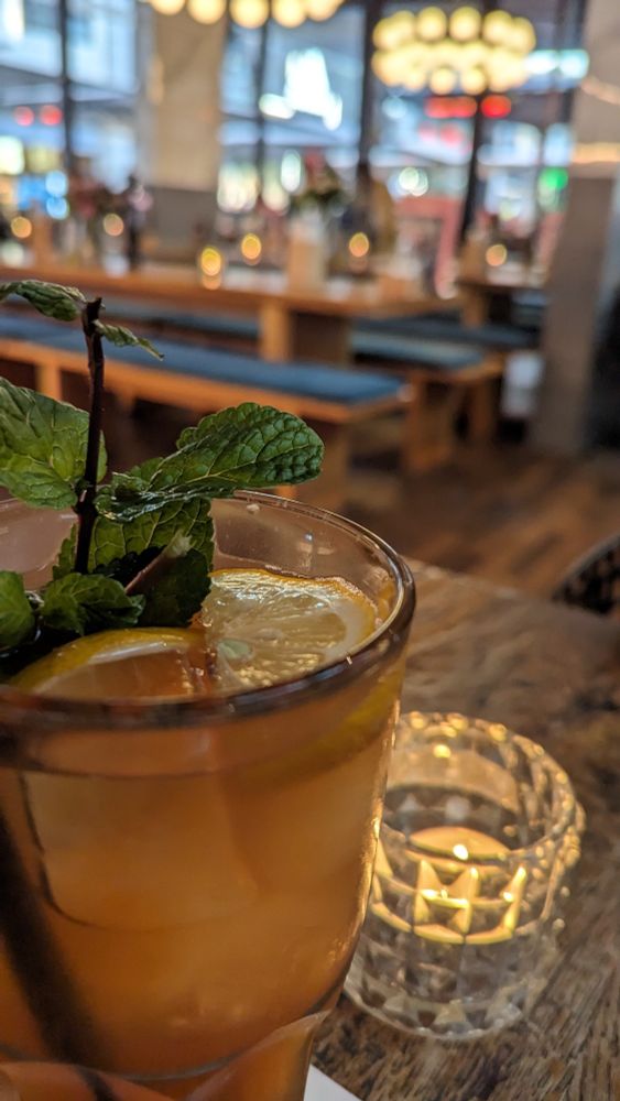 Ein Foto von einem Glas Zitroneneistee mit Eiswürfeln, einer Zitronenscheibe und Minze. Daneben steht ein Teelicht und im Hintergrund die Tische und Bänke des Restaurants.