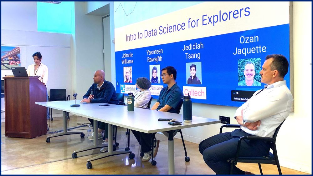 a panel of 5 people, with 1 standing at a podium and 4 others in chairs and with their names on a slide displayed on the wall behind them
