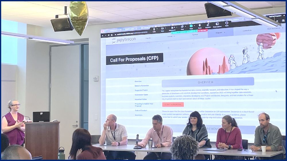 a panel of 5 people in chairs, with the moderator standing at a podium and with a slide displayed on the wall behind them featuring JupyterCon and a call for proposals