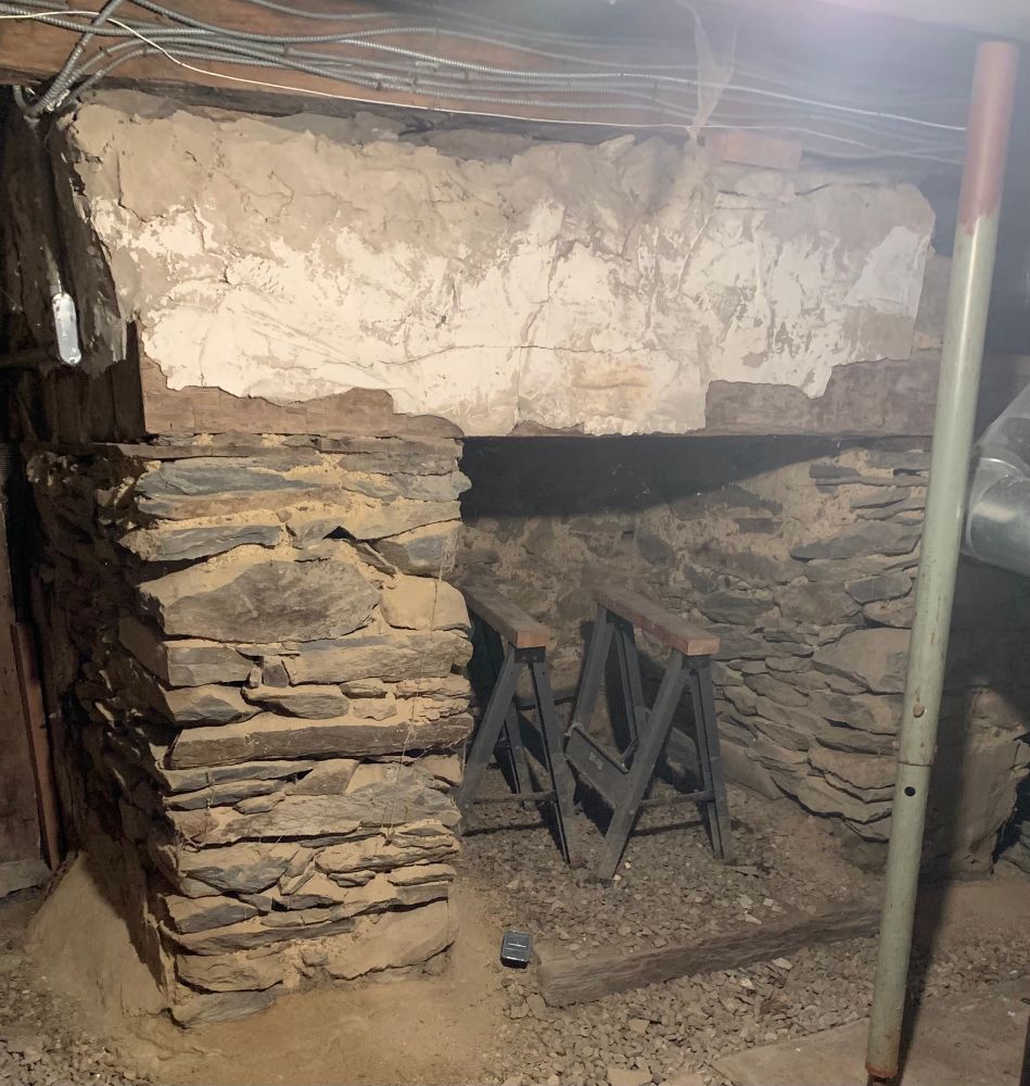 Large fieldstone fireplace in a basement with some sawhorses in it and a pole in front. 