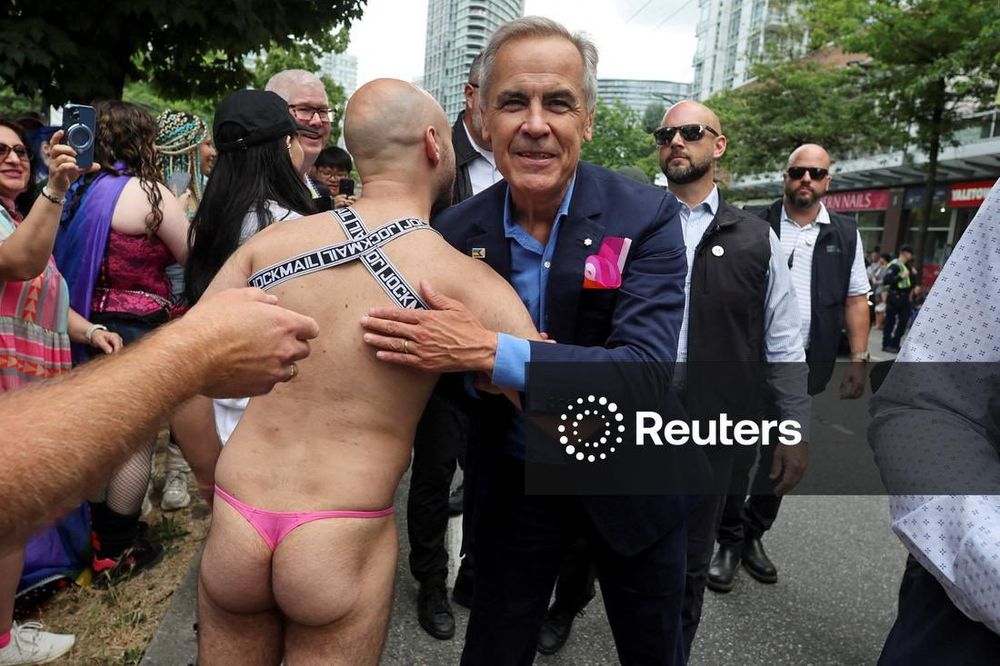 A man wearing a pink thong and harness interacts with another man in formal attire during a lively street event