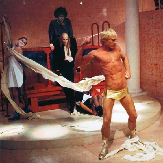 Rocky Horror Picture Show - A muscular man in golden shorts performs dramatically in a surreal scene with draped fabrics, surrounded by other figures in striking outfits.