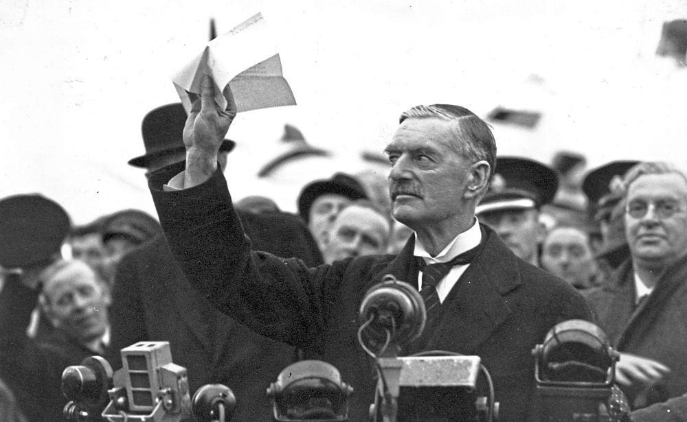 British statesman and prime minister Neville Chamberlain gestures with a paper in hand, addressing a crowd with vintage cameras in the foreground, capturing a historic moment.

(Central Press/Getty Images