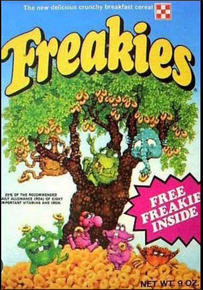 Image of a cereal box from the 1970s. Cereal is called “Freakies” and all the … are they mascots? I guess? Anyway the freakies depicted are 7 different weird little guys. One is red and looks like a grouchy crab another is kind of orange with lots of tentacles and he’s clinging to the tree branches (the box cover decent is a tree growing out of a hill made up of the Cheerios-like cereal, and there are 3 freakies in the tree and 4 on the hill underneath). There are 2 pink colored guys; one is rotund with a wide clam-like head, and another that’s similar to a dodo bird except just a proboscis like gonzo from the muppets. There’s a blue one up in the tree that seems like he’s all face, with no ears but a trunk like an elephant. And there are 2 green guys, one of whom looks kinda like a frog, but much more round? And another guy that’s just a round green dancing lump. Yes, they’re odd, if not actually attaining true freakishness. 