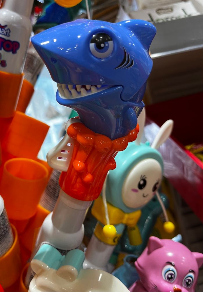 This is a closeup of one of a whole display of, well, I’m pretty sure they’re not just kids toys but also involve some kind of candy surprise inside the handle? This particular one is a shark, with a wily grin on his face, and he’s medium blue and he’s not quite winking — but he definitely looks like he’s up to no good. There’s some sort of trigger mechanism on the handle so he might have a bitey-jaw kind of thing going on too. For reasons that are not clear, the handle that he is emerging from looks like a melted candle, all wax dripping looking? But here’s the confusing part: it’s bright red— I’m not sure Sid they mean to make him emerge from waves and then just make a bad color choice? Or is he actually emerging from a big melt candle? I have no answers. 