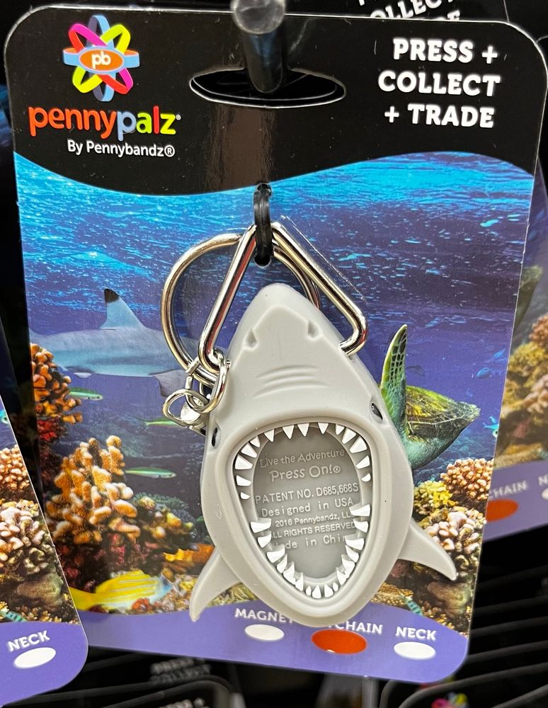 So this is a little rubber or silicone thing on a keychain that is designed to hold one of those pennies that you’ve put through one of those mechanical machines that rolls and stamps the penny into a design that makes it a souvenir of some destination. A national park, a museum, a famous place, or in this case, so help me, a Buc-ees store. This thing is called a “pennypalz” and the one in the pic is shaped like a shark whose jaws are wide open and you’re meant to put that souvenir penny into its mouth. Like a frame made of teeth. Delightfully ridiculous. 