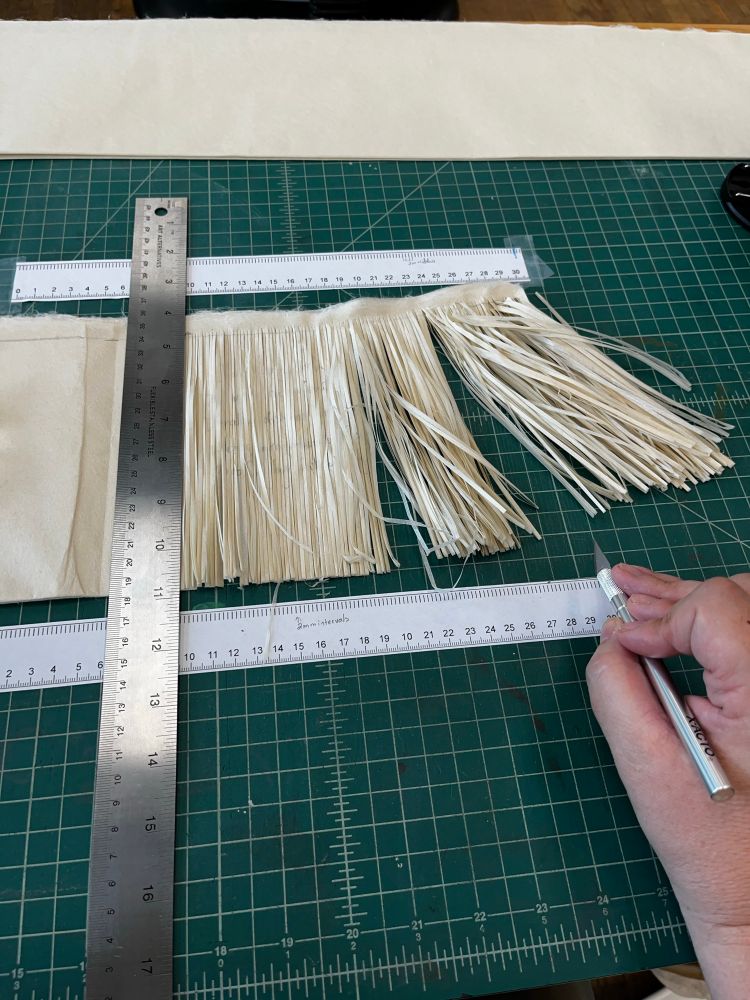 Cutting mat, green, with white grid marks and measurements. On the right is a hand holding an X-acto blade knife, because of what is center frame: a folded piece of Japanese kozo paper, set between two measuring tapes laid horizontally (above and below the paper) to guide the slicing of the paper into 2mm strips, by cutting along a steel ruler that runs vertically between matching points on the horizontal rulers. 