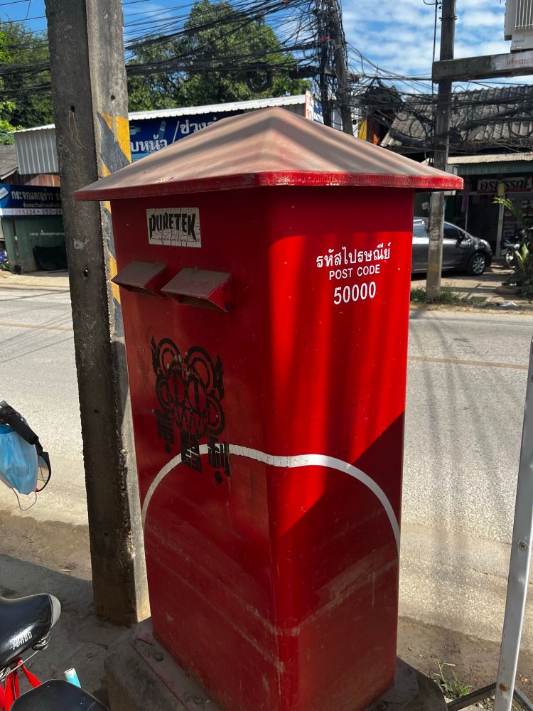 A red, but graffiti’ed with black spray paint, Thai Post column or pillar type postbox in Chiang Mai in post code area 50000. 