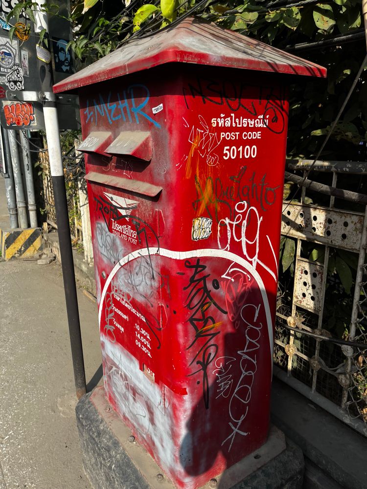 A red, but graffiti’ed with white and black spray paint, Thai Post column or pillar type postbox in Chiang Mai in post code area 50100. It has two deposit slots, and the collection hours are visible below. 