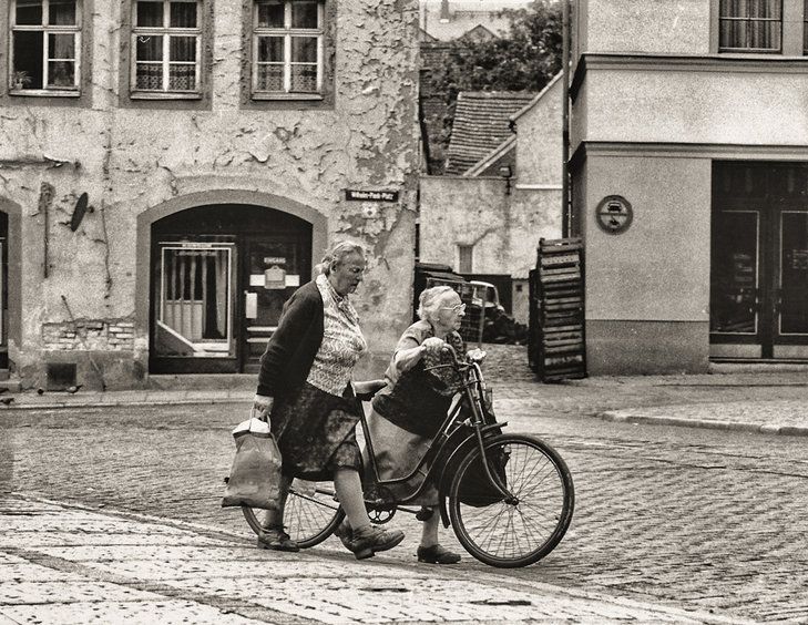 Black and white photo. Quiet town street (cobbled) with old buildings that have beauty and character despite their peeling paint and plaster. 2 elderly ladies, one pushing a bike that looks too big for her, both carrying bulging shopping bags, both striding out and looking very determined. 