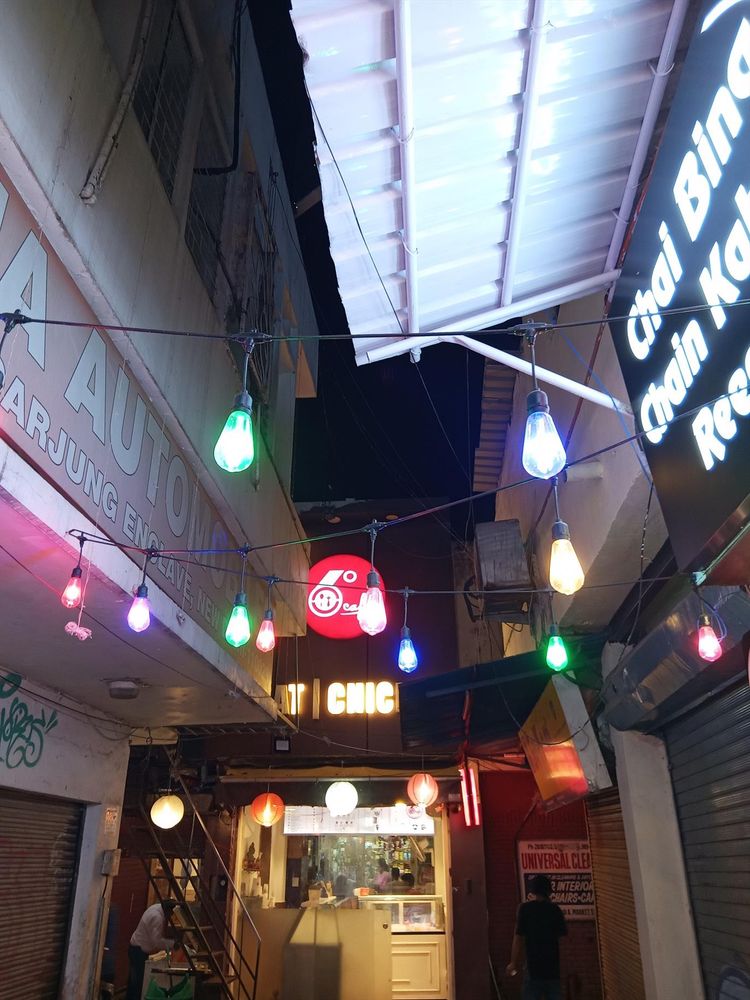 Picture of a narrow alley with hundreds of tiny lamps and neon signs. Under a tin-roof sky, waiters hand out food, while others offer haircuts and color printouts.