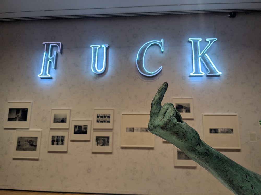 Copper cast of Ai Weiwei's arm flipping the bird, with neon letters spelling FUCK in the backdrop with some of his photography pieces of when he was in China 