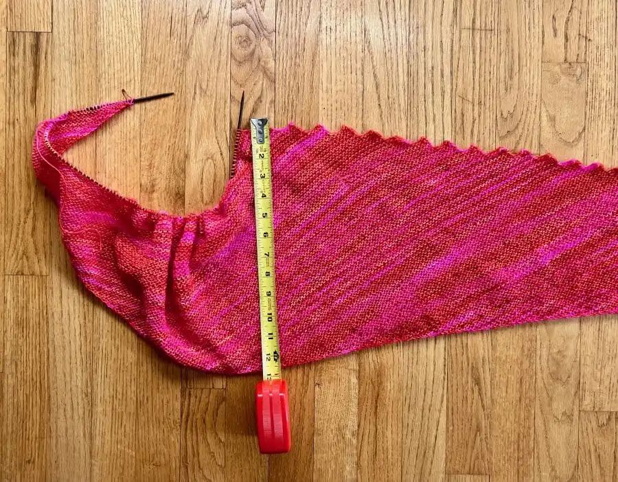 Crescent shaped shawl with saw-tooth edging in the process of being knit with a tape measure showing it is 13” tall. Color is a bright orange and pink variegated. 