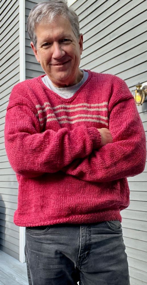 QueerJoe standing on his grey deck with his arms crossed, wearing a new shallow v-neck pullover in a color called Rose Heather with four narrow light tan stripes across the chest.