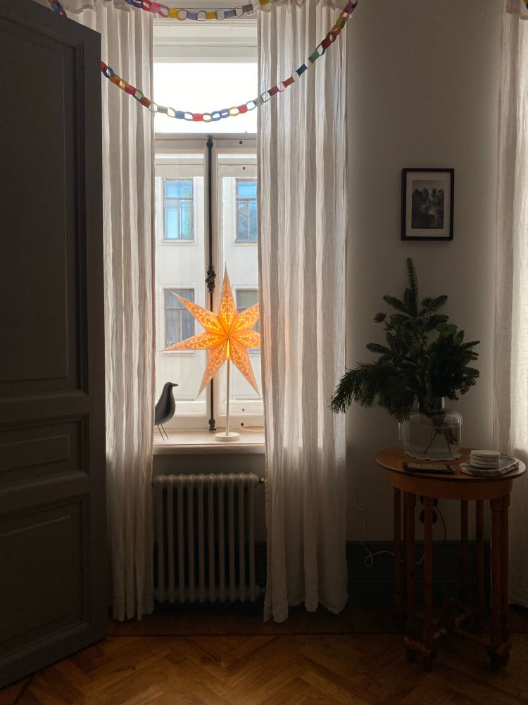 a photo of the window with wooden bird and paper star lamp on it. a table with a pine leaves next to it 