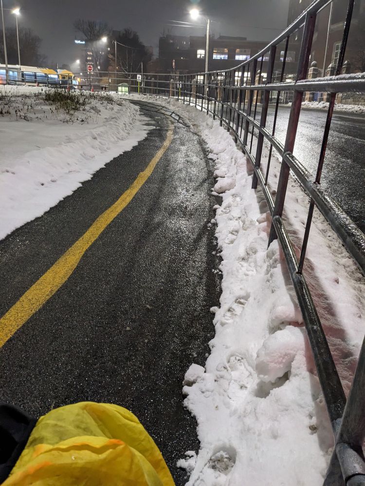 A partially cleared bike path with a yellow line showing. There are footprints on the snow on the right side. 