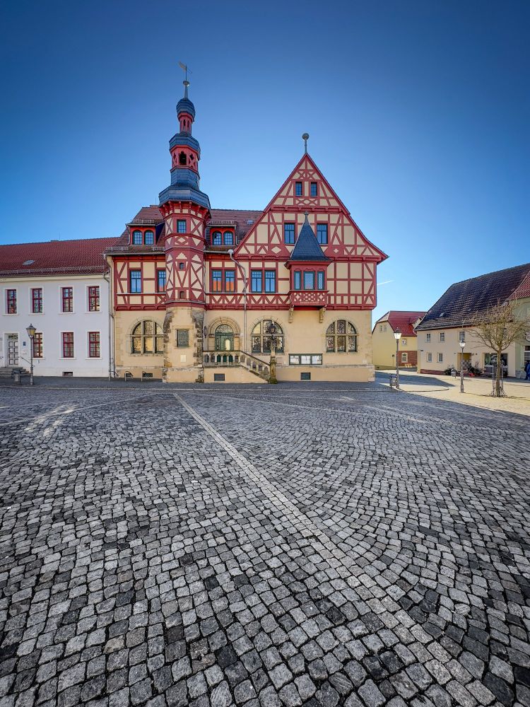 Das bearbeitete Bild zeigt das Rathaus in Harzgerode mit seiner markanten rot-weißen Fassade. Es ist ein historisches Gebäude mit einem hohen Satteldach, dekorativen Holzbalken und einem spitzen Turm mit einer Wetterfahne. Das Gebäude steht auf einem gepflasterten Platz, der mit unregelmäßigen Kopfsteinen ausgelegt ist. Die klare, sonnige Wetterlage und der blaue Himmel verleihen dem Foto eine frische und ruhige Atmosphäre.

(Bildbeschreibung aus GPT4)