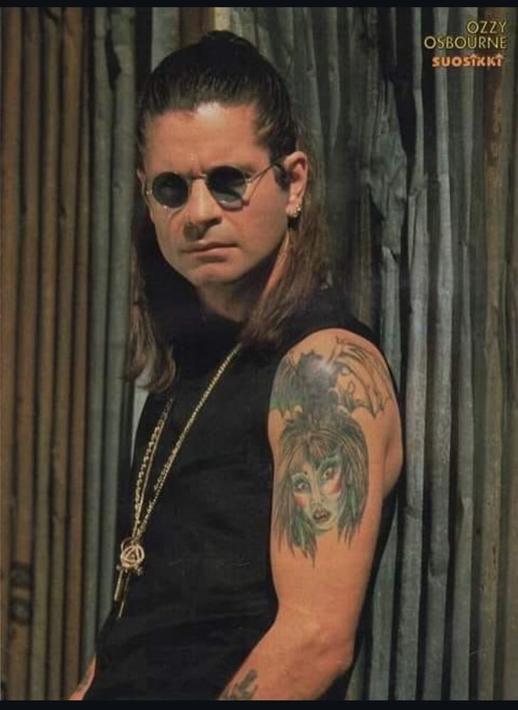 Foto do cantor Ozzy Osbourne encostado numa parede feita com um Metalon ondulado prateado. Ozzy está olhando para câmera, de óculos escuros e seu cabelo está com a metade superior presa em um coque alto. Ele usa uma camiseta preta sem mangas deixando as tatuagens do braço esquerdo a mostr, um colar longo, dourado com vários pingentes e o tradicional óculos escuros redondo com lente pequena. Ele está sério.