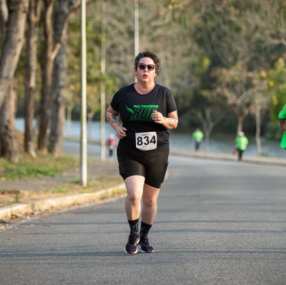 Imagem de uma mulher branca, cabelos curtos ecastanhos, de óculos escuros, batom vermelho e vestida com bermuda, meias, tênis e camiseta pretas, com o numero 834 preso na camiseta, correndo numa rua com arvores e pessoas desfocadas ao fundo.