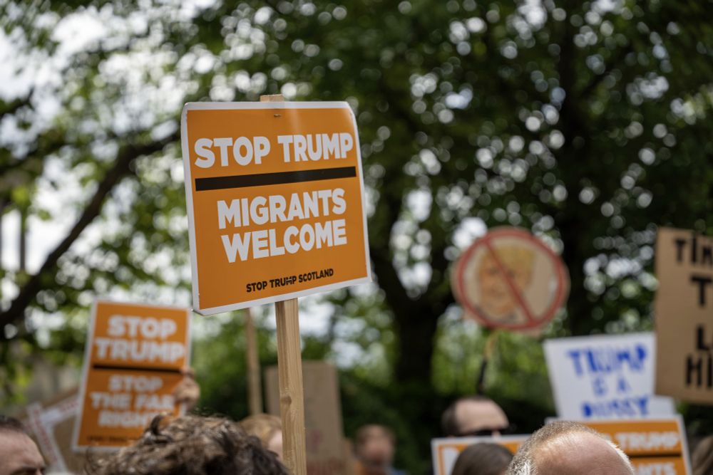 A sign held above a crowd reads 'Stop Trump, Migrants Welcome'.
